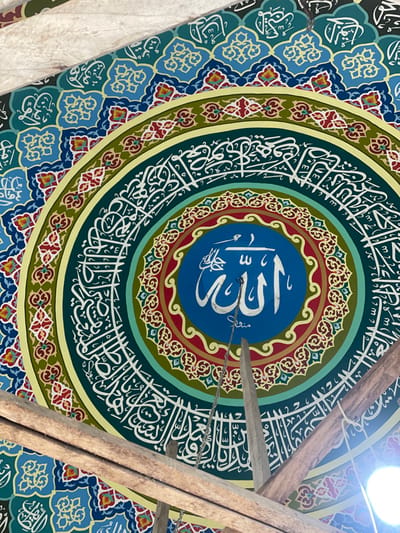 This image captures an upward view of an intricately decorated circular ceiling or dome interior, likely within a mosque in Kumbang, Indonesia. The central feature is a dark blue circular medallion, approximately 1/3 the width of the frame, displaying the word "Allah" in prominent white Arabic calligraphy. Below this, the word "Skrini" is written in smaller white script. A thin metal chain descends vertically from the center of the medallion.

The central medallion is surrounded by a series of concentric rings. Immediately outward is a band of red with yellow/gold floral patterns, followed by a wide band of teal with dense white Arabic calligraphy. A narrow yellow border separates this from another wide band of dark green, also filled with white Arabic calligraphy. The outermost visible band consists of blue sections patterned with light blue, yellow, and red geometric and floral motifs.

Three unpainted, raw wooden beams cross the lower portion of the frame, partially obscuring the decoration. One beam runs diagonally from the lower left towards the upper right. Another is horizontally positioned in the lower left corner. A third beam is visible vertically in the lower-center right, connecting to the diagonal beam. A bright, circular artificial light source is partially visible in the lower right corner, casting a glare. No individuals or specific actions are depicted.