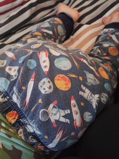 The image provides a close-up, high-angle view of a baby or young child lying down, focusing primarily on their lower body. The child is wearing blue pajamas or trousers with a detailed space-themed pattern. This pattern includes multiple rockets, some white with red accents and visible flames, astronauts in white suits, various planets and moons in shades of orange, red, blue, green, and grey with textured surfaces, and small yellow stars. The fabric of the clothing is visibly soft and shows natural creasing.

The child's bare feet are visible towards the top of the frame, resting on the surface beneath. This surface is covered by bedding featuring horizontal stripes in alternating colors of light brown/beige, dark green/black, and red. On the bottom left, a section of green camouflage-patterned fabric is visible, possibly another blanket or clothing item. A solid dark, likely black, fabric borders the scene on the bottom right. The overall setting is an indoor environment, most likely a bedroom, depicting a moment of rest or sleep. No specific time of day or weather conditions are discernible, nor are any emotions, as no faces are visible. There is no visible text in the image. The location context provided for this media is Teykovo, Russia.