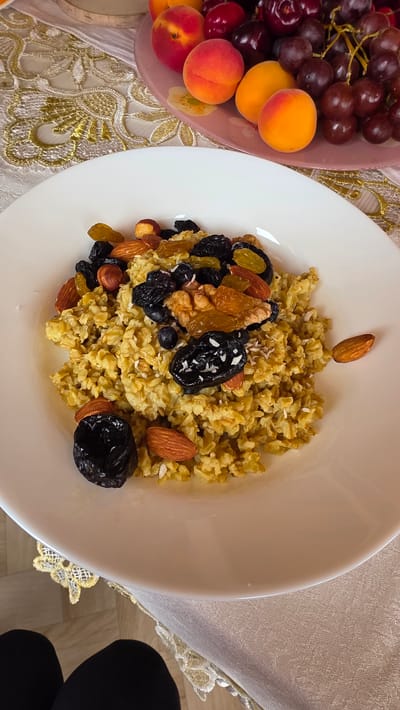 A high-angle, close-up shot captures a white bowl filled with oatmeal topped with various dried fruits and nuts. The oatmeal has a grainy texture and is a light brown color. Scattered across the top are plump, dark raisins, chopped almonds, a few whole hazelnuts, blueberries, and two large, dark prunes, one of which is sprinkled with shredded coconut. A single almond rests on the rim of the bowl. In the background, out of focus, is a pinkish-purple plate holding apricots and dark red grapes. The setting appears to be a table covered with a light-colored tablecloth featuring a gold embroidered pattern. The bottom of the frame shows the black leggings of a person seated at the table. The image does not contain any explicit information to determine the location within the city of Kupoyarovo, Russia.