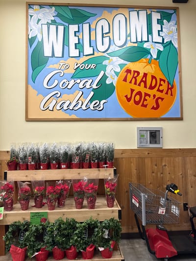 A vibrant Trader Joe's sign welcomes customers to Coral Gables with an orange illustration and floral details. Adjacent is a wooden display showcasing holiday plants. Rows of snowy fir trees, red poinsettias, and ivy wreaths in red wrapping paper pots are arranged on the shelves. A gray shopping cart, partially visible, sits next to the display, positioned against a wooden-paneled wall. A small, gray control panel is mounted on the wall between the sign and the plant display.