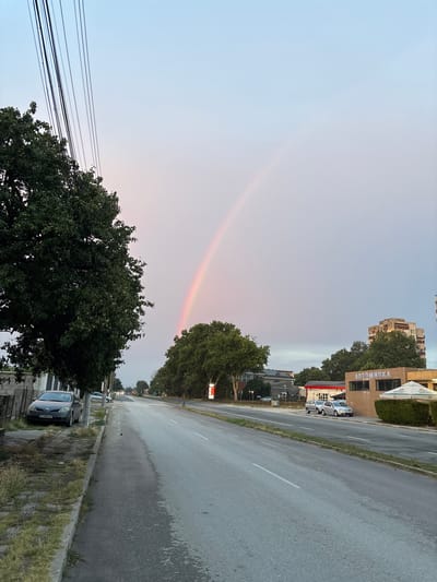 The image captures a view of a street in Aidemir, Bulgaria, with a faint rainbow arching across the hazy sky. On the left side of the street, a large tree with dense green foliage dominates the foreground. A dark-colored car is parked on the side of the road. The street itself is paved and appears wet in places, suggesting recent rain. Further down the road, trees line the right side, and buildings, including one with the Cyrillic sign "АВТОМИВКА" (Avtomivka, meaning car wash), are visible. The sky is overcast with a gradient of light blue and pink hues, creating a soft, diffused lighting condition. No people are clearly visible in the image.