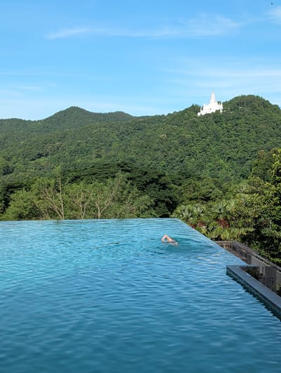 The image depicts an infinity pool with a person swimming in it, set against a backdrop of lush green mountains and a clear blue sky. The pool water is a vibrant turquoise, with visible ripples on its surface. A person, only their legs and lower torso visible, is swimming in the pool. The pool appears to be situated on a high vantage point, overlooking a dense, verdant mountain range. A white, ornate structure, possibly a temple or a monument, is visible on the side of one of the mountains. Palm trees and other tropical foliage are interspersed with the dense green of the mountains. The overall scene suggests a serene and picturesque tropical location. The time of day appears to be daytime, with bright sunlight creating a clear and vivid atmosphere. The weather seems pleasant, indicated by the clear blue sky. The location is identified as Ban Pong Nuea, Thailand. There is no visible text in the image.