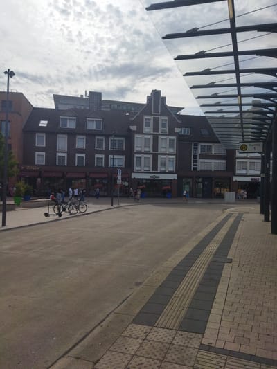 The image depicts a street scene in Monheim am Rhein, Germany, possibly near a bus stop indicated by a sign with "Bussteig 1". In the foreground, a paved area with tactile paving for visually impaired individuals leads towards a street or plaza. The middle ground shows a sidewalk with bicycles and an electric scooter parked. Behind the sidewalk, a row of buildings with shops and cafes is visible. One prominent shop has a "Tchibo" sign. People are seen walking and sitting outside the establishments. The background features a cloudy sky and more buildings, including what appears to be a modern structure. A glass-roofed canopy or walkway extends into the scene from the right side. The overall environment suggests a public, urban space.