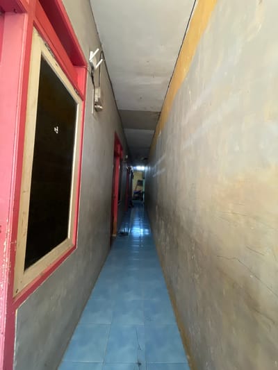 The image shows a narrow, tiled hallway within a building, likely a dormitory or apartment complex, in Anggaswangi, Indonesia. The hallway has blue tiled flooring and gray concrete walls. On the left side, there is a window with a pink frame. Above the window, a white Wi-Fi router is mounted on the wall. The right wall of the hallway is painted in a pale yellow or beige color with some scuff marks and faint lines. The ceiling is white and appears somewhat aged. At the end of the hallway, there are doors and what appears to be some equipment or storage items, suggesting a living or operational area. The lighting is dim, creating shadows that emphasize the narrowness of the space.