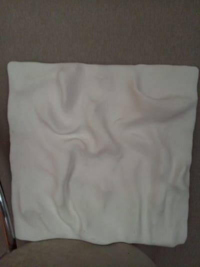 The image shows a close-up, overhead view of a white, rectangular object with a soft, pliable texture. The object is creased and wrinkled, suggesting it is made of fabric or a similar malleable material, such as dough or clay. The surface displays subtle undulations and shadows that create a sense of depth and contour. The object is placed on a textured surface that appears to be a brown fabric, possibly upholstery or carpet. A portion of a chrome chair leg is visible on the left side of the frame. The lighting is diffused, casting soft shadows across the object's surface. There is no indication of a specific location within the city of Vilyuysk, Russia, or any environmental context beyond the immediate foreground. No people or actions are depicted.