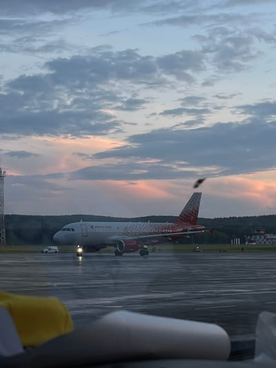 This media file captures a view of an airplane on a wet tarmac at dusk. The airplane, adorned with a distinctive red and white patterned livery, displays "РОССИЯ" in red lettering on its fuselage. Its engines are visible, and a red fin caps its tail. Several small vehicles are present on the tarmac, including what appears to be a white service vehicle. The tarmac itself is reflective, indicating recent rain. In the background, a line of trees separates the tarmac from buildings that resemble airport infrastructure, possibly control towers or terminals, with checkered flags visible on one structure. The sky is filled with dramatic clouds, tinged with the soft hues of sunset, suggesting a transition from day to night. The overall scene conveys a sense of a quiet, possibly post-rain, evening at an airport. The location is identified as Yemelyanovo Airport in Russia.