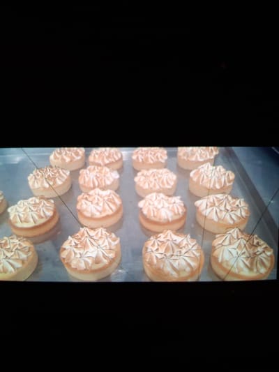This media file shows a close-up view of numerous baked goods, likely tarts or small cakes, arranged in rows. Each pastry is topped with a swirl of what appears to be meringue or whipped cream, artistically piped to create a textured, petal-like design. The base of the pastries is a golden-brown color, suggesting they are made of a cake or tart dough. The lighting in the image highlights the golden hues of the pastries and the creamy white of the topping. The scene is likely set in a bakery or kitchen environment, given the orderly presentation of the baked goods. The overall impression is one of freshly made, appetizing desserts ready for display or sale. No people are visible in the frame. The background is mostly dark, with the focus squarely on the pastries. The image quality is somewhat low, with visible grain and some digital artifacts, including lines across the pastries. There is no visible text in the image.