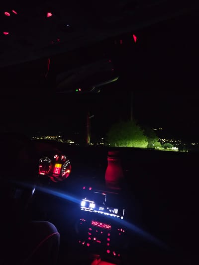 The image shows the interior of a car, likely at night. The dashboard lights and central console are illuminated. Outside the car window, there appears to be a view of greenery illuminated by streetlights. Given the prompt specifying Vratsa, Bulgaria, the location is likely within or near the city, possibly an area with streetlights and vegetation near a road. No people are clearly visible, although the presence of the car implies at least one occupant. The actions involve sitting in the car and looking through the window. The environment is dark outside, illuminated by street lights, and inside the car illuminated by the dashboard.