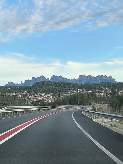 This photograph captures a scenic daytime view from a winding asphalt road, offering a glimpse of a Spanish landscape. In the foreground, the road curves gently to the right, prominently featuring a bold red and white striped median on the left lane and a solid white line on the right shoulder. Robust metal guardrails line both sides of the road, indicating its elevated position.
Beyond the road, the midground reveals a sprawling residential area or village nestled on a verdant hillside. Numerous houses and buildings with light-colored roofs are visible, interspersed among dense green trees and vegetation. The overall impression is that of a community integrated into the natural environment.
Dominating the background is the distinct and majestic silhouette of the Montserrat mountain range, characterized by its unique, jagged peaks and towering rock formations. The mountains rise dramatically against a bright blue sky, adorned with scattered white and light grey clouds. This iconic formation serves as a key geographical indicator, confirming the location context of Sant Salvador de Guardiola, Spain, which lies in the vicinity of Montserrat.
The image suggests a pleasant journey or drive through this picturesque region. The time of day appears to be late morning or early afternoon, with clear weather conditions. No specific human activity beyond implied travel is depicted, and no visible text is present in the scene.