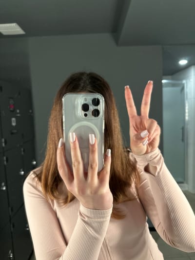A person is taking a mirror selfie in a locker room. The person is holding a clear phone case with a dark-colored phone inside. They are wearing a light pink, ribbed long-sleeved shirt and have light-colored, square-shaped nails. They are holding up two fingers in a peace sign. The background shows dark gray lockers on the left and a light blue shower stall on the right. The floor appears to be tiled. The image does not contain any specific visual cues that would allow for the identification of the city of Carlow, Ireland.