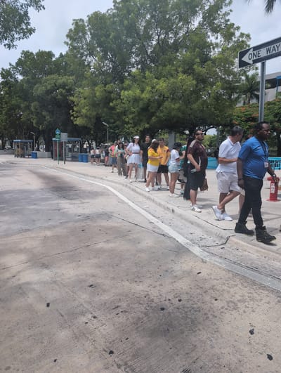 The image captures a queue of people standing on a concrete sidewalk next to a street. The people are varied in age and gender, dressed in casual summer clothing. In the background, there are trees, a "ONE WAY" street sign, and what appears to be a bus stop with blue trash receptacles. The environment suggests an urban setting, possibly during the day given the lighting. A few individuals in the foreground are walking, while others are stationary in the line. One person is wearing a blue shirt and black pants, another a white polo shirt and shorts, and a woman in a white dress is also visible. The pavement shows signs of wear and tear, with some dark stains and tire marks. The overall scene depicts a public area where people are waiting, possibly for transportation or an event. The city of Gladeview, United States is mentioned as the location.