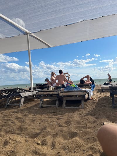 The image depicts a beach scene under a canopy. In the foreground, sand is visible with several wooden lounge chairs, some occupied by people. In the background, the sea stretches out under a blue sky with scattered clouds. Several individuals are seen on the beach and in the water. The location is identified as Gres-2, Kazakhstan.