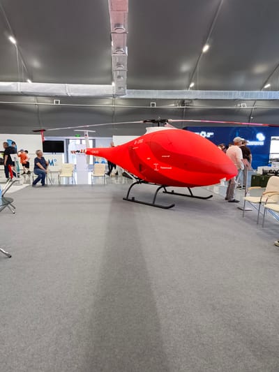 This is a photograph of a bright red drone, a helicopter-like unmanned aerial vehicle, displayed indoors on a grey carpeted floor. The drone has a large, bulbous fuselage with prominent lettering that appears to be in Cyrillic script. It is mounted on a black skid landing gear. In the background, there are several people observing the drone and other exhibits. The environment appears to be a convention or exhibition hall, with a high ceiling and visible ventilation ducts and lighting fixtures. The overall impression is of a modern technology showcase. The text "B-200" is visible on the drone's side. Another visible text reads "ЗАПРЫТО" and "ОТКРЫТО" indicating 'closed' and 'open' respectively. Further text visible is "АЭРОДРОМ" and "КАНИНСКИЙ". The location is stated to be Korsakov, Russia.