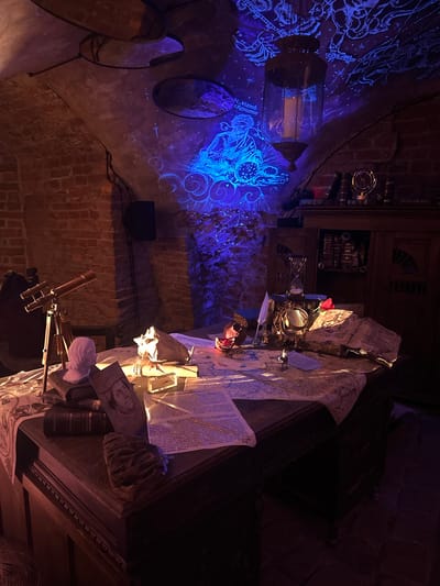 This image depicts a dimly lit, atmospheric room in Guryevsk, Russia, designed to evoke an ancient study, alchemist's lab, or astronomer's observatory. The room features exposed brick walls and an arched ceiling, creating a historic and intimate setting.
The left wall and ceiling are bathed in a vibrant blue and purple light, projecting intricate patterns of celestial maps, constellations, and swirling cosmic designs, creating a magical ambiance. Several oval-shaped mirrors are affixed to this wall, reflecting the luminous projections. A dark, ornate lantern-like object hangs in this section, contributing to the antique feel.
Dominating the right side is a heavily laden wooden desk or table, piled with an array of historical objects. Numerous open books and rolled-up papers, some with detailed illustrations and visible text, are scattered across the surface. A portrait of a person is discernible on one of the documents. Scientific instruments include a brass-colored telescope in the foreground, an hourglass on the desk, and a compass-like device mounted on the wall above. A single red rose adds a splash of color to the collection, which also features a skull-like object and other various ornate items. The desk itself boasts carved details, and shelves filled with books are visible in the dark background, further emphasizing a theme of intellectual pursuit. A dark speaker is mounted on the brick wall to the left of the desk.
The artificial lighting suggests a nocturnal setting, and the overall arrangement evokes a sense of mystery, wonder, and historical exploration. No people are present, indicating the scene is either a display or a meticulously arranged space for contemplation.
Visible text on the documents includes "artin Zuber.", "M.Mamlı sRomanus", "Der Riber", "Ons Chriftue", "bette", "Goner", "me guac", "facb/", "rom", and "ber".