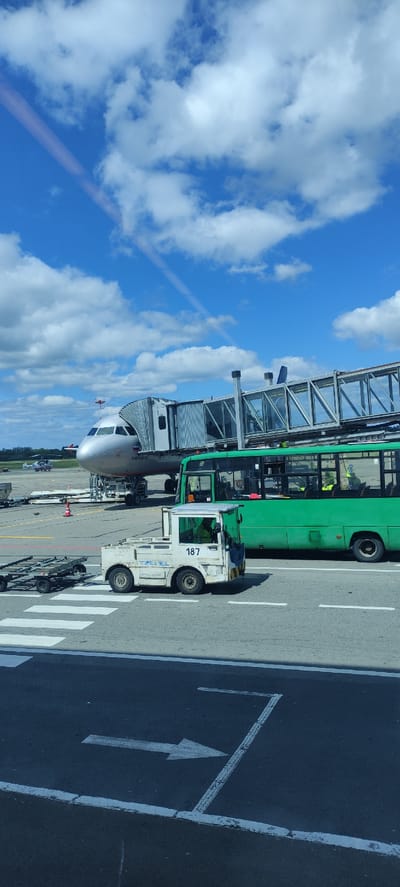 The image captures a vibrant daytime scene at an airport tarmac, specifically at Khrabrovo, Russia. A passenger jet, likely an Airbus A320 or similar narrow-body aircraft, is prominently featured, with its nose and front fuselage visible. It is connected to a modern glass-paneled jet bridge, suggesting it is either preparing for departure or has just arrived.

To the right of the jet bridge, a long, green airport bus is parked, its windows reflecting the bright sky. Adjacent to the bus and closer to the foreground, a white airport ground support vehicle, possibly a baggage tug or pushback tractor, is positioned. This vehicle clearly displays the number "187" on its side. Below this number, some additional, less legible text can be seen.

The ground features clear white markings, including a pedestrian zebra crossing and directional arrows painted on the dark asphalt, indicating movement patterns on the apron. The sky above is a brilliant blue, dotted with fluffy white clouds, and a faint contrail is visible diagonally across the upper left quadrant, hinting at other aircraft activity. The scene is well-lit, suggesting it is midday under clear conditions.