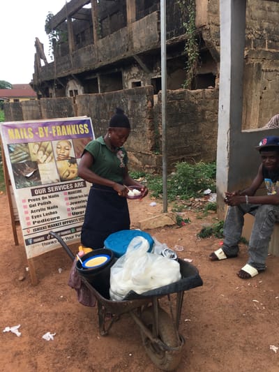 This image captures a lively street scene in Isieke, Nigeria, showing two individuals and a mobile nail salon setup. The setting is outdoors, characterized by reddish-brown earth and a weathered, multi-story concrete and brick structure in the background, partially overgrown with green vines, suggesting an older building or perhaps one under construction/deconstruction.
In the foreground, a woman, dressed in a green polo shirt and dark skirt with a black head covering, is seated on what appears to be a modified wheelbarrow or cart. She holds a small bowl containing white contents, seemingly engaged in an activity related to her nail services or possibly preparing food. Beside her, a large banner stands prominently, advertising "NAILS-BY-FRANKISS." The sign lists "OUR SERVICES ARE" Manicure, Press On Nails, Gel Polish, Different Nails, Acrylic Nails, Jeweleries, Eyes Lashes, Training, Pedicure, and Upgrade. It also includes contact information with social media handles and the address "ISHIEKE MARKET OPPOSITE IZZI," explicitly confirming the location.
Above and slightly behind the woman, a young man wearing a cap, a graphic t-shirt, and patterned jeans sits on a concrete ledge. He looks down towards the woman and the cart with a slight smile, his hands clasped. The ground around the cart shows some discarded items, typical of a bustling market area. The time of day is daytime, indicated by the natural light, and the overall atmosphere appears active and informal.