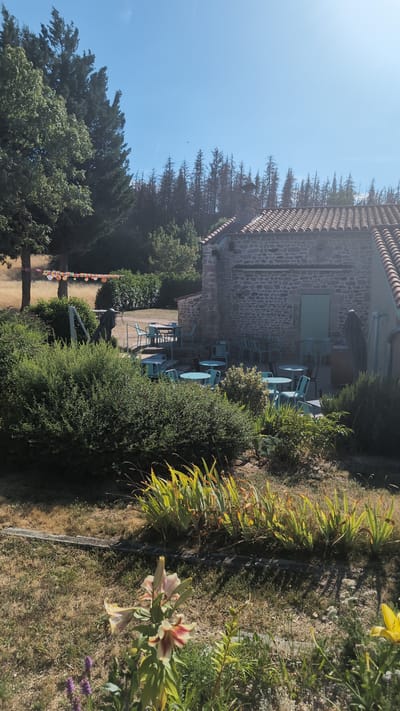 The image depicts an outdoor seating area of a building constructed from stone, likely a restaurant or cafe, in Chambles, France. The building has a tiled roof and a light green door. Several turquoise tables and chairs are arranged on a patio area. In the foreground, there are lush green bushes and various flowering plants, including lilies. Behind the building, a dense forest is visible under a clear blue sky. The overall scene suggests a pleasant, sunny day.