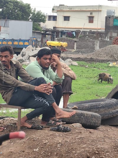 In Padli, India, three men are seated on a bench by a construction site. The man on the left wears a dark olive shirt and black pants, his legs crossed. The man in the center wears a light green shirt and black pants, with his legs crossed. The man on the right wears a brown shirt and dark pants. A pig with mottled brown and black coloring is visible in the background, foraging in a grassy area with piles of dirt and construction materials. Behind the men are stacks of large stones. In the distance, a light-colored building with a brown trim and several windows is visible, along with other structures including corrugated metal buildings and a blue truck with yellow lettering. Two large tires are stacked in the foreground on the right side of the image.