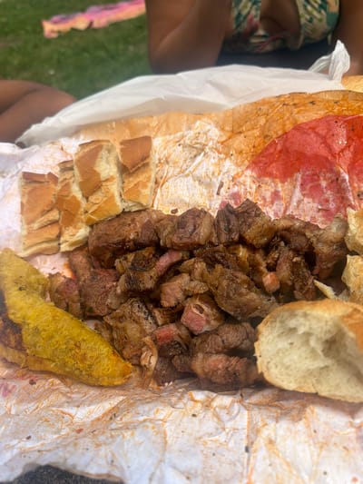 A close-up, slightly elevated view shows a picnic spread on a grassy area. In the foreground, a pile of meat, appearing to be steak or beef, is presented on a crumpled piece of white paper. Sliced bread pieces are arranged to the left and right of the meat. A piece of fried plantain, a yellowish-green elongated item, is positioned to the left of the meat. In the background, out of focus, is a person's legs and torso, wearing a floral patterned top. Another person's legs are visible on the far left, and a pink and yellow blanket or cloth is on the grass behind them. The environment suggests an outdoor setting, possibly a park or picnic area. There is no visible indication of the city of Campezo/Kanpezu, Spain in the image.