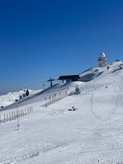 This image displays a snow-covered mountain ski slope on Mount Jahorina, Bosnia and Herzegovina, under a clear blue sky. In the upper right quadrant, a white building featuring a spherical dome, consistent with a weather station or observatory, is situated on the mountain peak. Below and to the left of this structure, a dark-colored ski lift terminal building is visible. A ski lift tower supports cables extending from this terminal, with multiple ski lift chairs suspended along the line.
The mid-ground and foreground are extensively covered in snow, crisscrossed by numerous ski tracks. Approximately fifteen individuals, identifiable as skiers, are dispersed across the slope; some appear to be actively skiing downwards, while others are standing stationary. A long, multi-section snow fence, constructed of wooden slats, extends diagonally from the mid-left towards the upper right, delineating a section of the slope. Another partial section of snow fence is visible in the lower-mid ground. Several small, snow-laden evergreen trees are sparsely distributed across the terrain. In the mid-ground right, a low, geometric object, partially covered by snow, casts a shadow. The scene is brightly illuminated by direct sunlight, indicated by distinct shadows cast by all prominent objects and structures.