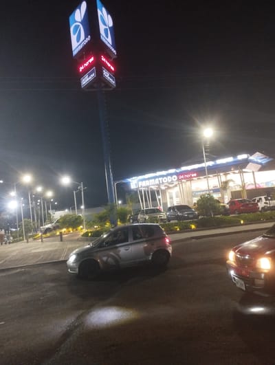 The image captures a nighttime scene in what appears to be a parking lot in front of a "Farmatodo" store, a pharmacy chain operating 24 hours. The store's sign is prominently displayed. A tall pole with the brand's logo and the "24 Horas" and "Auto Farmacia" is seen. Several cars are present, including a silver hatchback and a red car with headlights on. Streetlights illuminate the scene. There are some plants and trees planted in a median. The OCR confirms the presence of a "Farmatodo" and "24 horas" text. No people are clearly visible. Given the logo and the text found by the OCR, and the specific architecture, it's possible this is the Farmatodo store in Barquisimeto, Venezuela.