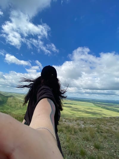 A first-person perspective image shows a person with long, dark, wind-blown hair and a black baseball cap from behind, looking out over a vast, green, rolling landscape under a bright blue sky with scattered white clouds. The person is wearing a black t-shirt and has tattoos on their arm, including a silver chain bracelet on their wrist. The foreground is rocky and covered in dry grass, transitioning to smoother, verdant hills and fields in the middle ground, with a distant line of trees and the horizon visible. The scene appears to be a natural outdoor location, possibly a hilltop or viewpoint. The image does not provide any specific identifiers for the city of Andreevka, Russia.