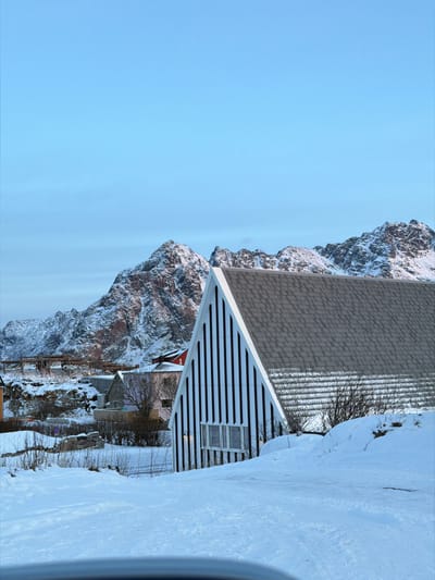 The image depicts a winter scene in Henningsvær, Norway. The foreground consists of snow-covered ground, exhibiting gentle undulations. In the midground, a prominent building with a steeply pitched, triangular facade and a dark grey shingle roof is visible. The visible side of this building features distinct vertical black and white stripes and a horizontal row of rectangular windows. Snow partially covers sections of the roof and is accumulated around the base of the structure. To the left and behind this building, several other structures, some appearing residential, are scattered across snow-covered terrain. Elevated to the left, scaffolding or similar construction elements are present. The background features rugged, snow-capped mountains under a clear, light blue sky. Sparse, bare vegetation is visible in various locations, largely obscured by snow. No individuals, animals, or explicit actions or interactions are present. A curved, dark grey surface is partially visible at the bottom edge of the frame, suggesting the photograph was taken from within a vehicle.