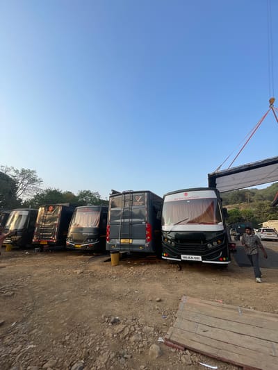 The image depicts a roadside scene in Mira-Bhayander, India. A line of dark-colored buses, potentially vanity vans for a film shoot, are parked on a gravelly area. Overhead, a rigging system with cables and a large, white rectangular panel is suspended, possibly for lighting or diffusion. A man wearing a plaid shirt and jeans stands near the lead bus. The sky is clear and blue. A stack of wooden planks lies on the right side of the image. License plates are visible on the buses.