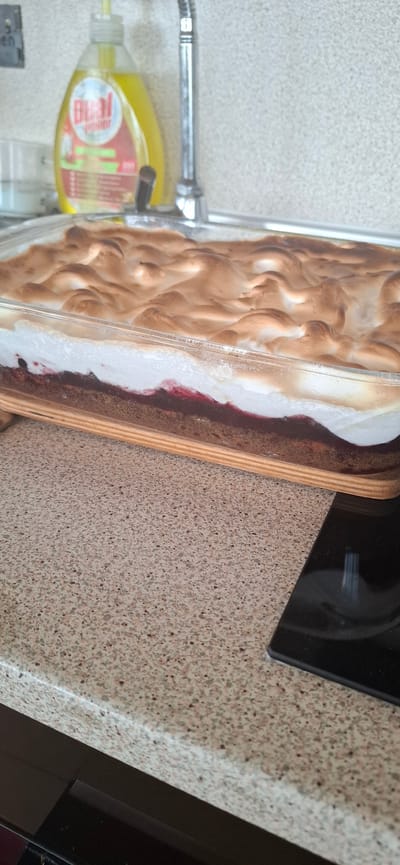 A baked dessert, likely a cake with a meringue topping, is presented in a clear rectangular glass baking dish. The dessert has distinct layers: a dark base, a layer of dark red fruit filling, and a thick, browned meringue that has peaks and browning. The baking dish rests on a speckled countertop. In the background, a yellow bottle of dish soap and a chrome faucet are visible, suggesting a kitchen environment. The image is taken from a medium-high angle, focusing on the dessert. The scene is domestic and suggests food preparation or presentation in a home. The specific location in Dobrinishte, Bulgaria, is not visually evident from the image itself, which depicts a generic kitchen setting.