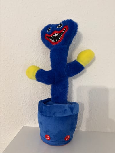 A plush toy resembling the character Huggy Wuggy from the game Poppy Playtime stands in a blue pot on a white shelf against a textured white wall. The toy is predominantly blue with a large, toothy grin, red lips, and black and white eyes. It has yellow-tipped hands. The blue pot has two small red circular buttons with musical notes on them. This scene is located in Velbert, Germany.