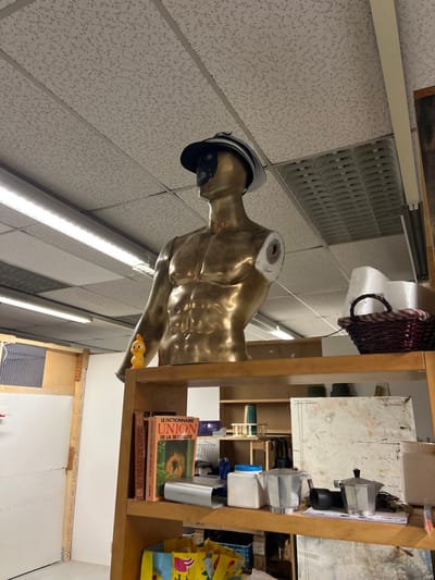 A headless golden male torso wearing a black hat stands beneath a ceiling with vent and fluorescent lighting. Beside the mannequin is a wooden shelving unit stocked with books, including "Le Dictionnaire Union de la Sexualite", storage containers, and coffee makers. A yellow rubber ducky clings to the edge of the mannequin. The room's background includes visible sections of a white wall, window, and wooden beam.