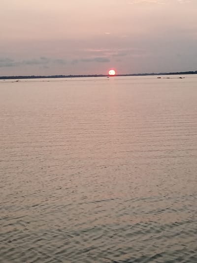 The image displays a wide-angle view of a large body of water under a twilight sky, identified as being in the city of Akoko, Nigeria. The dominant feature is the expansive water surface, exhibiting gentle ripples that create a textured, reflective appearance, primarily in soft grey-pink and muted blue tones. A prominent, bright, reddish-orange sun, partially submerged or very low on the horizon, is visible slightly right of the center in the upper third of the frame. Its shape appears somewhat flattened horizontally. Above the sun, the sky transitions from pale pink and peach hues near the horizon to a lighter grey-pink higher up, with faint, elongated cloud formations present. A thin, dark horizontal line, likely distant land or dense vegetation, demarcates the horizon across the entire width of the image. Several indistinct, small landmasses or clusters of vegetation are scattered on the water's surface near the distant horizon, particularly towards the right side. No people, specific man-made objects, or observable actions are present in the scene.