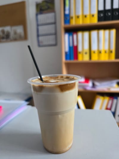 A close-up, eye-level shot shows a plastic cup filled with a layered coffee drink and a black straw inserted into it. The drink has a lighter base and a darker, frothy layer on top. The cup is positioned on a light gray surface, which appears to be a desk or table. In the background, a bookshelf filled with colorful binders is visible. To the left of the bookshelf, a corkboard with some framed pictures is mounted on the wall, and a calendar with multiple rows of text is also present. The scene suggests an indoor office or study environment. There are no people or animals present in the image. The location is in Gara Oreshets, Bulgaria.