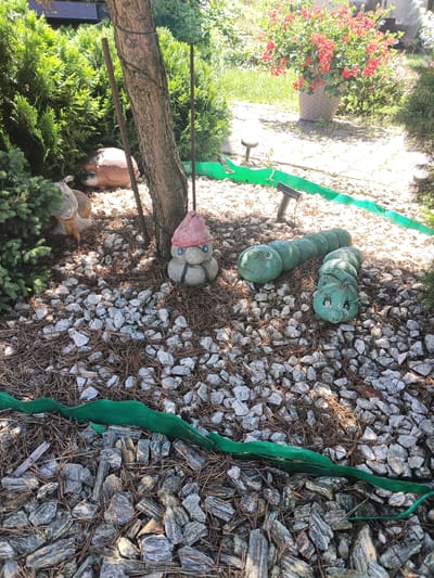 The image depicts a garden scene in Igołomia, Poland, likely during daytime with sunny weather, given the bright lighting and shadows. The foreground is covered with a mixture of small grey stones, larger grey and blackish rocks, and fallen pine needles. A bright green border, possibly a garden edging, snakes through the rocky ground.

The primary subjects are garden ornaments. To the left, nestled among green shrubs, is a small, light brown garden ornament resembling a hedgehog or a similar small animal, with painted facial features. Next to it is another ornament, a gnome-like figure with a pink hat, blue eyes, and a black outline for a mouth. In the center and to the right, a segmented green caterpillar ornament lies on the rocks, its body composed of several round sections and its head featuring painted eyes and a mouth. Behind the gnome and caterpillar is the trunk of a tree, with a rough, textured bark, and two dark metal rods are embedded in the ground near it.

In the background, more lush green bushes and a large pot overflowing with bright red flowers are visible. The overall impression is a cheerfully decorated garden. There is no visible text in the image.