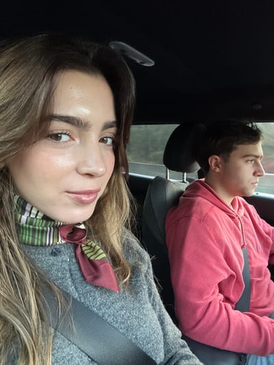 The image displays two individuals occupying the front passenger and driver's seats of a vehicle. A young adult female is positioned in the foreground, occupying the passenger seat. She has long brown hair, a clear complexion, and is looking directly at the camera with a slight smile. She wears a grey knit garment and a patterned silk scarf around her neck, featuring stripes of green, white, pinkish-red, and brown, with a solid reddish-brown section. A black seatbelt diagonally crosses her torso. To her right, in the background, a young adult male is visible in profile, seated in what appears to be the driver's seat. He has short dark hair and is looking forward, out of frame to the right. He is dressed in a red, ribbed hooded sweatshirt and also has a black seatbelt across his chest. The car interior features a dark headliner and dark grey upholstery. Through the side windows, blurred green foliage is visible, suggesting movement through an area with natural vegetation, such as a road or highway. The specific location within Leiria, Portugal, is not discernible from the visual evidence provided in the image. The ambient lighting indicates a daytime setting with soft illumination.