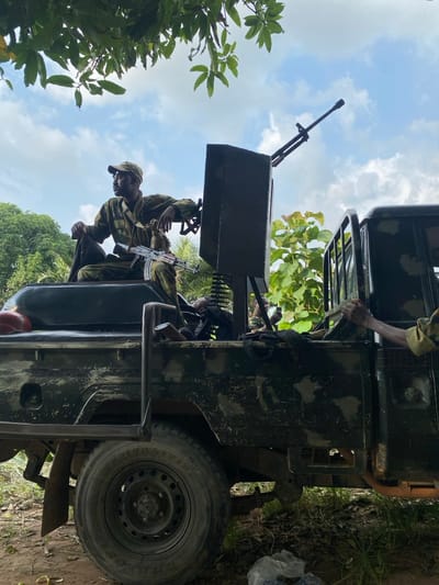 The image depicts a dark green utility truck, possibly a pickup, in a vegetated outdoor setting under a partly cloudy sky in Ayati, Nigeria. A male individual, wearing camouflage fatigues and a cap, is seated on the roof of the truck's cab, holding an AK-variant assault rifle across his lap, looking to the viewer's left. A heavy machine gun, mounted on a custom dark grey platform, is positioned in the truck bed, pointing upwards and to the right. At least two additional individuals, also in camouflage, are partially visible within the truck bed; one's right arm extends towards the truck's side railing, while another's head is visible behind the weapon mount. The truck's body is soiled, and a red object is present in the rear of the bed. Dense green foliage, including overhanging tree branches, frames the scene. The ground beneath the vehicle is unpaved and scattered with leaves, and a white plastic bag containing dark contents lies near the bottom right.