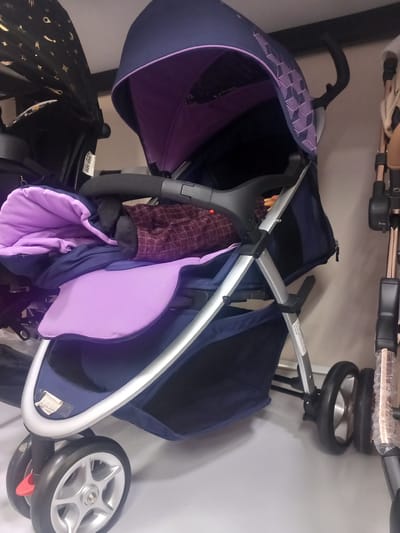 A dark blue and purple stroller is displayed in a retail environment, likely a store, in Khodja Akhrar Vali, Uzbekistan. The stroller is the primary subject and is positioned in the center of the frame. It features a dark blue frame, a dark blue canopy with a purple interior, and a purple seat pad. A dark fabric blanket or cover is draped over the seat. The stroller has multiple wheels, with the front left wheel clearly visible and showing a silver five-spoke design. The background consists of a plain light-colored wall and the partial views of other strollers, suggesting a commercial setting. A white sticker with black text is attached to the stroller's frame. The lighting appears to be artificial and general store lighting. There are no people present in the image.