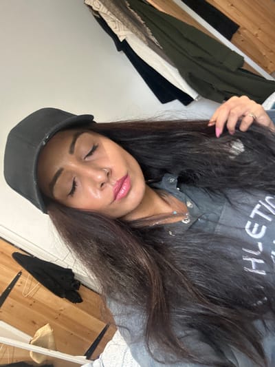 An indoor portrait features a young woman with long, dark brown hair, captured in a selfie or close-up shot that is rotated 90 degrees clockwise. Her eyes are closed, and her lips are slightly parted with glossy pink lip makeup. She has a small silver nose ring and wears multiple delicate necklaces, one with a small blue bead. Her right hand, showing light pink manicured nails and a ring, rests near her head.
She is dressed in a dark grey long-sleeved top with white lettering, partially visible as "HLETIC," and a black baseball cap. The setting appears to be a casual domestic space, likely a bedroom or dressing area in Winslow, United Kingdom. In the background, various clothing items are visible, including a dark green garment, a cream-colored lace-trimmed item, and black fabric, suggesting clothes hanging or draped over furniture. Wooden furniture, possibly shelves or a dresser, frames the left and right sides of the image, along with a light-colored woven item, possibly a straw bag or hat, on the right. The overall mood is relaxed and peaceful.