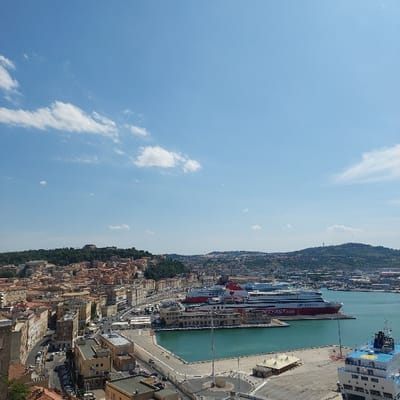 This image captures an elevated panoramic view of the city and port of Ancona, Italy, under a clear blue sky with sparse white cumulus clouds. The foreground and midground are dominated by the port's turquoise water basin and surrounding infrastructure.

Two large, red and white passenger/car ferries, prominently branded "SUPERFAST.com" and "ANEK LINES," are docked along the western pier of the port. A section of a third vessel, blue and white in color, is visible in the lower right corner of the frame, partially extending out of view. The port features concrete piers, dockside buildings, and a circular roundabout area. A light-colored, canopy-like structure is situated on one of the piers.

Adjacent to the port, the city of Ancona unfolds with densely packed multi-story buildings, characterized by light-colored facades and red or terracotta tiled roofs. Numerous vehicles are visible on the streets and parked throughout the urban area. The city extends up the surrounding hills, which are covered in green vegetation. A prominent historical structure, possibly a fortress, is discernible on a distant hilltop to the left. No individual persons or specific human interactions are discernible due to the elevated perspective.