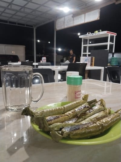 The image depicts a close-up view of a table at an outdoor eatery in Sigli, Indonesia, at night. In the foreground, a clear glass mug filled with water sits on a reflective surface. To its right, a bright green plate holds several cylindrical food items wrapped in what appears to be banana leaves and grilled, with some charring visible. Next to the food, a small clear plastic container with a green lid contains a light-colored powder, and a pink label with the word "SHIN" is affixed to it. In the background, a person is visible standing in front of a white shelf unit. The overall environment suggests a casual dining setting under a covered area, with artificial lighting illuminating the scene.