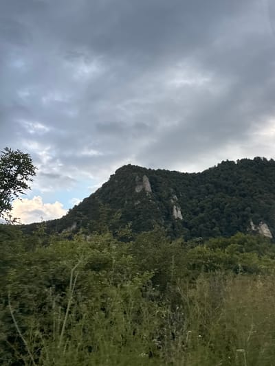 The image depicts a mountainous landscape under a cloudy sky. A steep, tree-covered mountain face dominates the right side of the frame, with some rocky outcrops visible. The vegetation appears dense and lush. On the left side, the sky is filled with dramatic, dark gray clouds, suggesting overcast or impending stormy weather. Patches of lighter sky and some brighter clouds break through the darkness, hinting at a possible break in the weather or sunlight filtering through. In the foreground, the branches of a leafy tree are visible, adding a sense of depth and framing the scene. The overall impression is one of a wild and somewhat moody natural environment. The time of day is unclear due to the overcast conditions, but it appears to be daytime. There is no visible text in the image. The location is identified as Voronov, Russia.