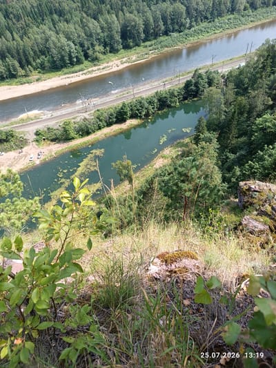 The image shows a panoramic view of a wide, winding river bordered by dense green forest. On the left bank, a sandy beach is visible with a few small boats or kayaks in the water. A railway track runs along the river's edge. On the right bank, a narrower, darker body of water is connected to the main river, with patches of aquatic vegetation. The foreground features a rocky cliffside covered with dry grass and a few leafy plants. The scene appears to be a natural landscape with some man-made infrastructure. The timestamp "25.07.2026 13:19" is superimposed on the bottom right corner. The image location is stated to be in Brishtamak, Russia.