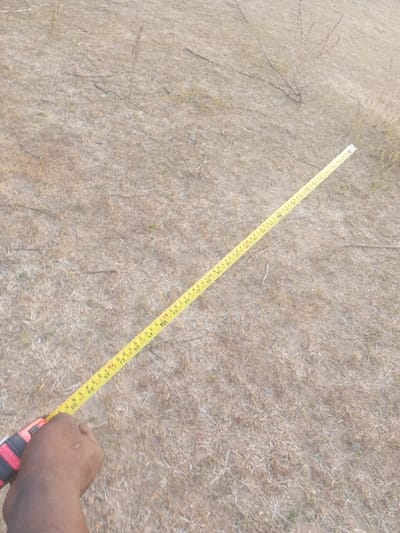 The image displays a forensic overhead view of a dark-skinned human hand holding and extending a yellow measuring tape across a dry, uncultivated outdoor area in Okeni, Nigeria. The hand, visible in the lower-left corner, grips the tape's red and black casing. The yellow tape extends diagonally towards the upper-right, featuring black numerical markings indicating measurements in both inches and feet (e.g., "70" and "2F"). The immediate environment consists of parched, light brown to tan earth covered with sparse, dry vegetation, including blades of grass and small, scattered twigs, characteristic of a fallow or arid landscape. No other individuals or significant structures are visible.