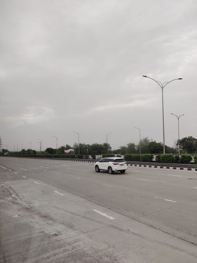 An exterior, eye-level, vertical shot captures a white SUV driving on a multi-lane highway under a cloudy sky. The SUV is positioned in the right lane, moving away from the viewer. The highway is paved with concrete and marked with white dashed lines. On the left side of the highway, a median strip with shrubs and black and white striped barriers separates it from the other side. In the distance, trees form a dense line along the edge of the highway. Several streetlights with multiple arms are visible along the roadside, some with lights on. A large truck with a red cab and a covered trailer is parked or moving slowly in the far distance on the other side of the highway. The overall atmosphere is overcast and subdued, with a sense of vastness due to the open road and the expansive sky.