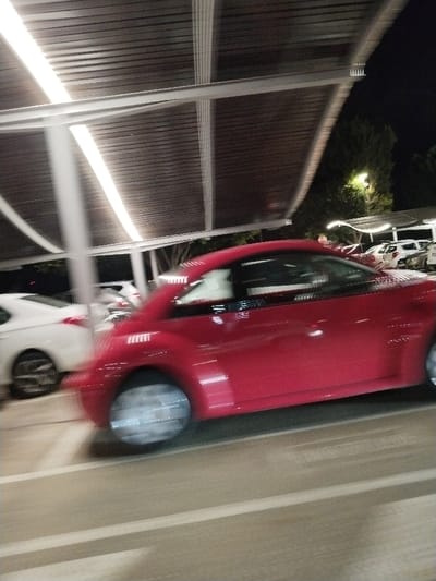 A red car drives through a parking garage at night. The lights above illuminate the corrugated roof. Other cars can be seen parked in the background, with trees and streetlights visible in the distance. Motion blur suggests the car is moving. White lines are visible on the parking lot surface.