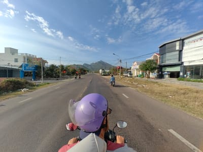 The image depicts a first-person perspective from the rear of a motorcycle traveling forward on a paved two-lane road in Tuy Phong 4, Vietnam. In the immediate foreground, the back of a person wearing a purple full-face helmet with a raised clear visor is visible, along with parts of the motorcycle's handlebars and mirrors. A conical hat (nón lá) rests behind the helmet.
The road ahead is asphalt, marked with a double white line separating the two lanes of traffic traveling in the same direction. Grassy shoulders line both sides of the road.
On the left side of the road, a white multi-story building with a blue sign is visible. Further down, additional buildings, power poles, and scattered vegetation, including small trees and shrubs, are present. A light-colored animal, possibly a dog, lies on the grassy shoulder near a patch of disturbed earth.
On the right side of the road, a modern multi-story commercial building with dark and white paneling and large windows is prominent. It features a partially visible sign with text that appears to include "KHOAN." Further down, smaller buildings, trees, and power poles are observed. The shoulder on this side consists of dry grass and scattered refuse.
Multiple motorcycles are visible ahead on the road. One individual in a blue shirt is riding a motorcycle in the right lane. Another two motorcycles are further ahead on the left side of the road. A white truck is also present further down the right side of the road.
In the distant background, a range of mountains rises under a clear blue sky with wispy white clouds. The overall scene suggests a sunny day in a semi-urban or rural area, characterized by local commercial activity and residential structures.