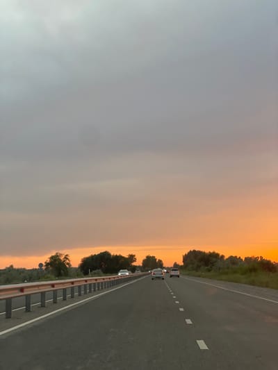 The image depicts a highway in a rural setting, likely during sunset or sunrise, given the warm orange and yellow hues in the sky. The sky is a dominant feature, transitioning from a hazy grey on the left to vibrant orange on the right, suggesting either the beginning or end of the day. A line of trees and vegetation borders the right side of the road, creating a natural backdrop.
The highway itself appears to be a two-lane road with a white dashed line marking the lanes. Several cars are visible, driving in the same direction as the camera. One car, a light-colored SUV or minivan, is in the foreground on the left lane, followed by other vehicles further down the road. A guardrail with reflective elements runs along the right side of the highway, separating the road from the embankment and vegetation.
The overall mood is serene and a bit melancholic, possibly due to the overcast sky on one side and the fading light on the other. The scene suggests a journey on a road trip or a commute through the countryside. There is no visible text in the image. The location context is Ivanovka, Kyrgyzstan, which aligns with the natural landscape depicted.