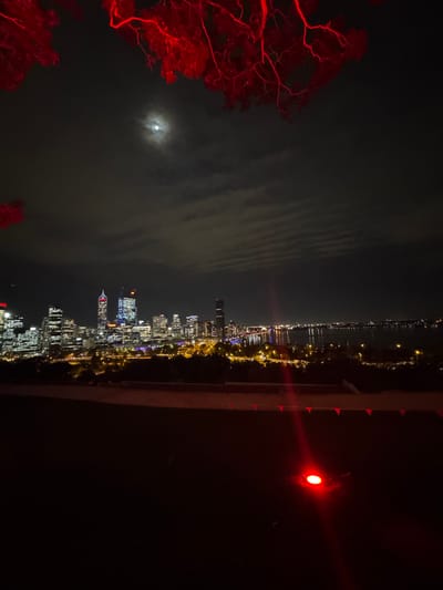 The image captures the Perth skyline at night, as viewed from Kings Park, Australia. The Swan River is visible, dotted with lights. The city's illuminated skyscrapers pierce the dark sky. The moon is partially obscured by clouds. In the foreground, red-lit trees frame the view. A red light is prominent on the right. The scene suggests a nighttime vantage point overlooking the cityscape.