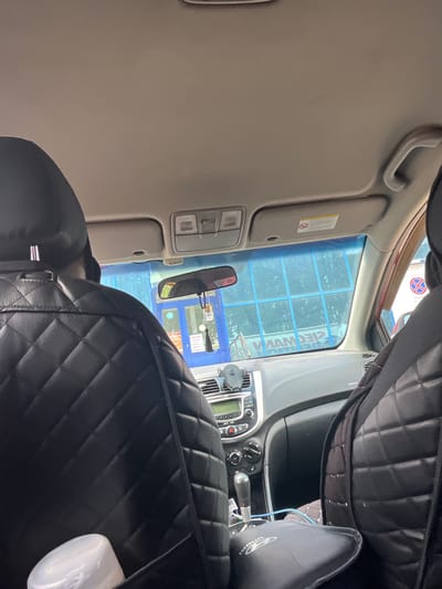 The image is taken from inside a car, looking forward through the windshield. The interior of the car features black, quilted seat covers. The dashboard is visible, with a car stereo and air conditioning controls. A phone mount is attached to the dashboard. Outside the car, a blue building with large windows is visible. There appears to be some sort of service area or establishment with signage in Cyrillic, one sign reads "СТОП" (STOP). The sky is visible through the tinted windshield. The scene is likely a street or parking lot in the city of Novaya Otradovka, Russia, possibly near a car service center.