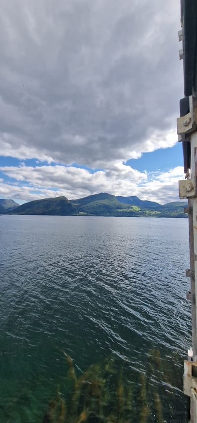 The image shows a wide shot of a body of water, likely a fjord, with a distant mountainous landscape under a partly cloudy sky. On the right side of the image, a portion of a metal structure, possibly part of a bridge or a ferry, is visible. The water has ripples and is a dark blue-green color. The distant mountains are covered in lush green vegetation, with some patches of lighter green that may indicate fields or clearings. The sky has a mix of dark grey clouds and bright blue patches, suggesting intermittent sunlight. There are no people visible in the image. The scene appears to be captured from a moving vessel or a fixed elevated point overlooking the water. The location is Volda, Norway, as indicated by the reference to the city in the prompt.
